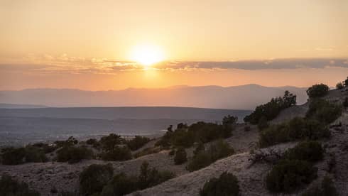 A hill with small trees with the sun setting behind it.