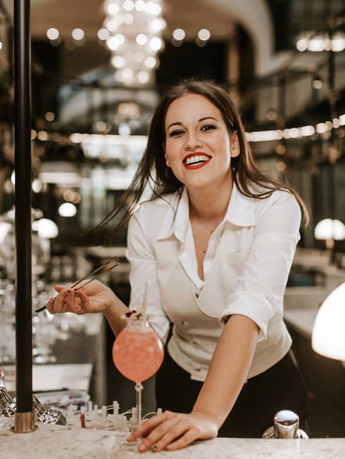 Bartender Anett Schurina in white blouse, smiling and leaning over a M�KVIR�G cocktail