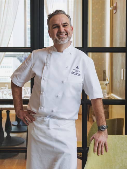 A male chef in white chef clothes standing in front of windows.