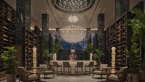 Chandelier Bar with six jade pillars flanked by wooden shelves and potted palms, glass-topped bar in the middle with six cocktail chairs, giant chandelier hanging overhead
