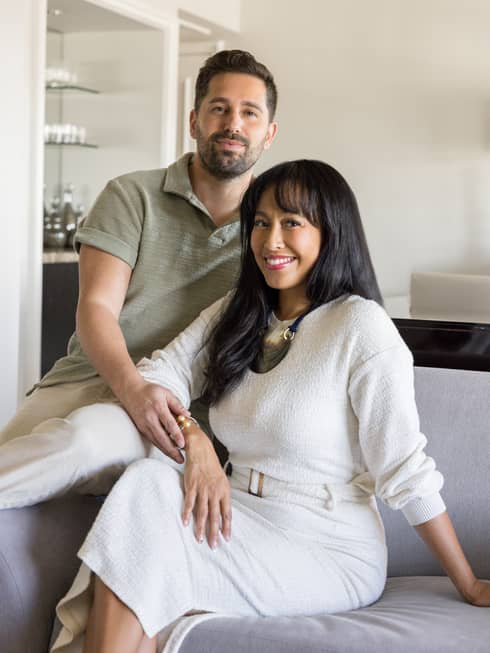 A man with short dark hair and a trimmed beard wearing a sage green polo shirt and khakis sits on the arm of a grey sofa next to a woman with long dark hair wearing an ivory sweater and maxi skirt