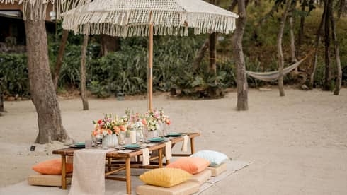 Wooden dining table set up with floral centrepieces and candles beneath an umbrella on a beach