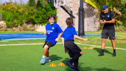 Soccer coach talking to two young guests exercising outside with a basketball court in the background.
