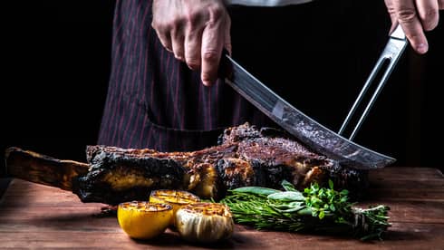 Close-up of chef slicing large rack of grilled meat, vegetables with butcher knives