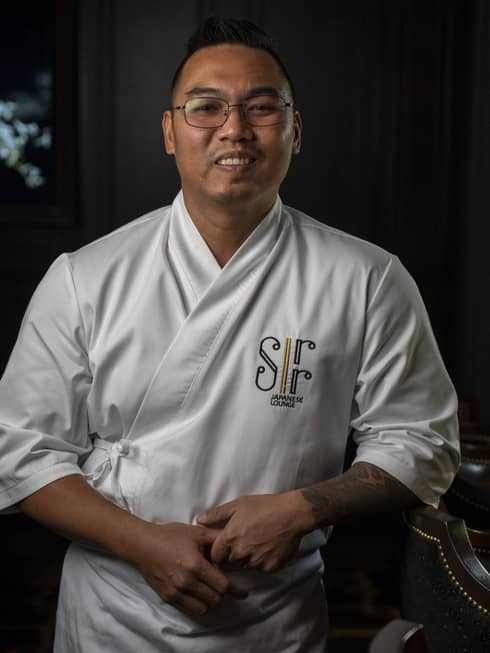 Chef with short dark hair and glasses, wearing a white chef's jacket and apron, stand smiling at the camera