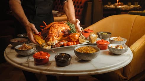 Server wearing black shirt places a whole roasted turkey on a white, round marble table also filled with small bowls of holiday dishes