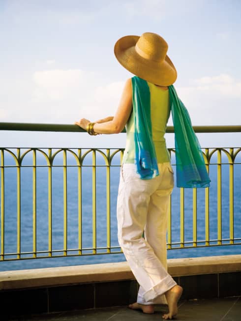 Back view of woman wearing sun hat, flowing blue-and-green scarf, standing at patio railing looking at sea
