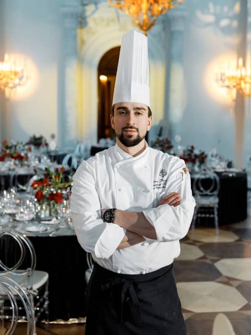 Chef Denis, dressed in a chefs hat and apron, stands in the middle of a restaurant with his arms crossed against his white chef's jacket..