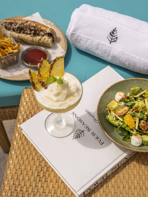 Two plates of food and a cocktail served on a teal lounge chair and small rattan table