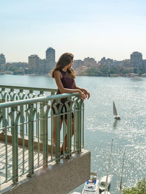 A woman standing on a balcony looking out at a large river below and a city in the background, there is a boat sailing the river.