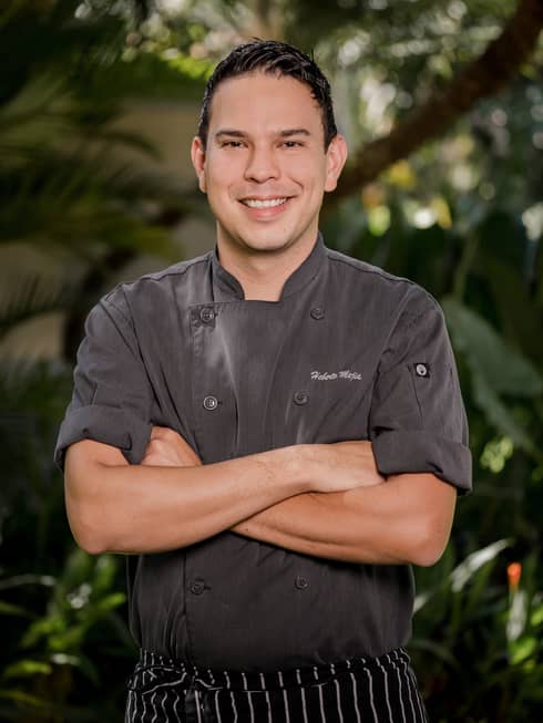 Chef Heberto Mejia ? wearing a black chefs coat with the sleeves rolled up ? stands smiling at the camera with is arms crossed