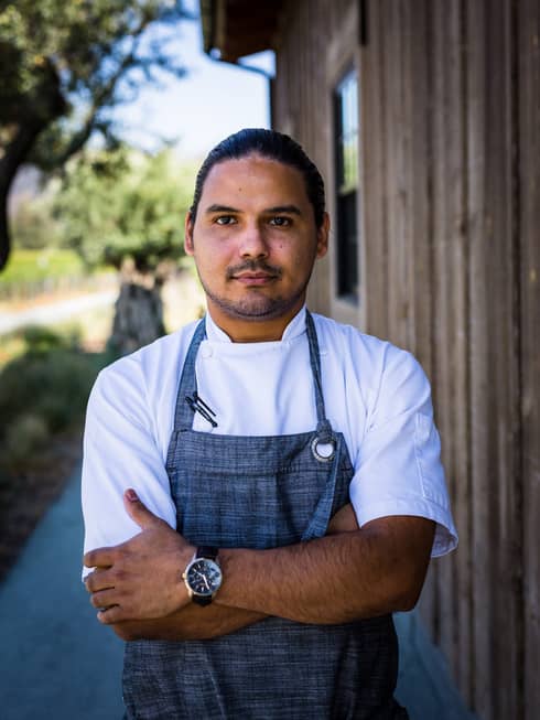 A chef in a blue apron standing outside near a wood building.