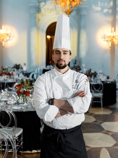 Chef Denis, dressed in a chefs hat and apron, stands in the middle of a restaurant with his arms crossed against his white chef's jacket..