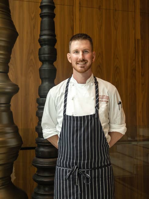A man in a pin-striped apron stands in front of an oak wall and banister.