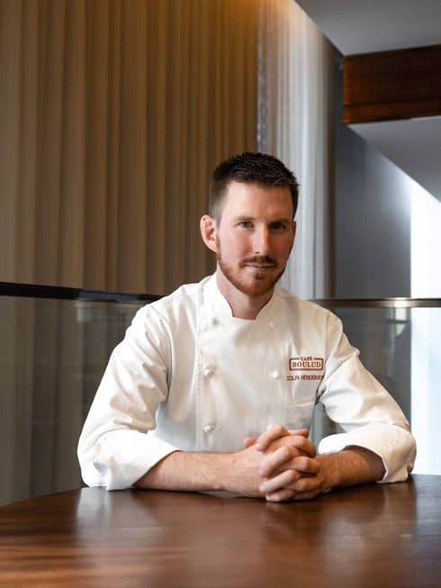Chef de Cuisine Colin Henderson sitting at a wooden table in his chef whites