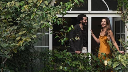 Two people stand on a vine-covered balcony surrounded by foliage.