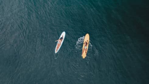 Aerial view of two guests lying on their stomachs on surfboards facing each other on calm water, each wearing a safety leash.