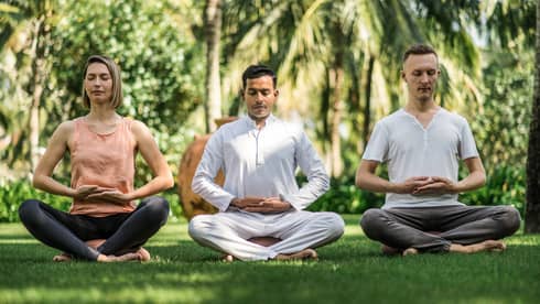 Three people meditating in a seated position on a grassy lawn, surrounded by tropical greenery, with their hands resting gently on their laps.
