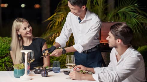 At a table for two amid foliage under the night’s sky, a server grates truffle over a diner’s dish as another diner looks on.