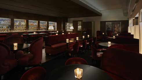 A dimly lit lounge with red chairs and a large selection of drinks behind a bar.