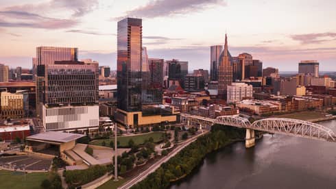 Aerial view of downtown Nashville, Tennessee at sunset, featuring modern skyscrapers, a river, a bridge, and a park with greenery in the foreground.