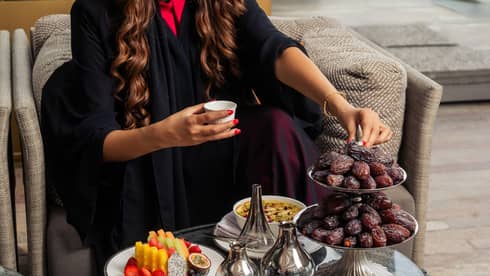 Person with long dark curly hair wearing a black full-length dress reaches for a date served on a two-tiered silver platter next to a silver tea set and two white plates of food