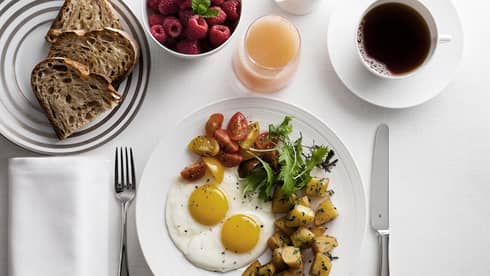 Breakfast plate with sunny-side-up eggs, cherry tomatoes, greens, and roasted potatoes, alongside toast, raspberries, grapefruit juice and coffee.