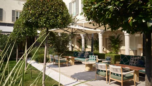 Exterior courtyard at Four Seasons Hotel Milano, with patio chairs, tables and umbrellas, grasses and manicured trees.