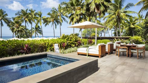 Luxury resort suite private terrace featuring a small rectangular pool, wooden lounge chairs with flush white cushions, and a dining table with palm tree ad ocean views