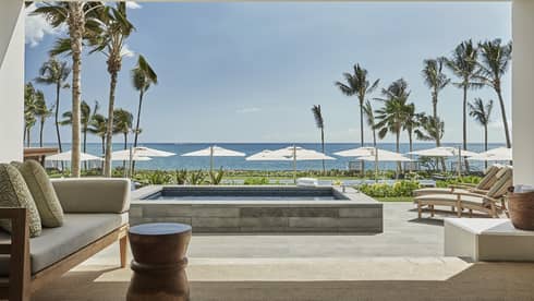 Pacific Suite's luxurious beachfront patio with a comfortable seating area, a private pool and sun loungers facing the ocean, framed by palm trees and umbrellas under a clear sky