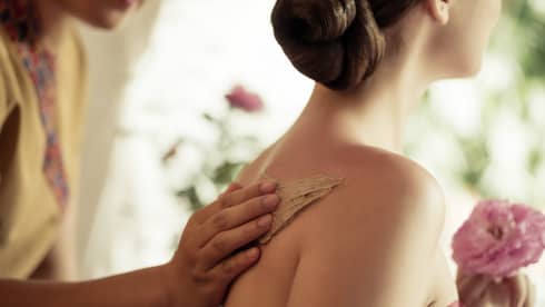 Spa staff rubs thick lotion on woman's bare shoulders as she holds a fresh flower