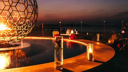 ,Night-time view of a table for two set next large round fire pit,Night-time view of a table for two set next large round fire pit with candles surrounding the border