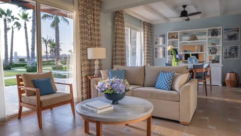 Guest room living area with tan sofa, armchair, round marble coffee table, dining table in background, sliding glass door to patio