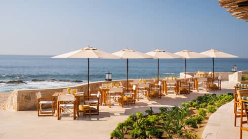 Open-air terrace with a row of tables and umbrellas set along a curved stone wall overlooking the ocean