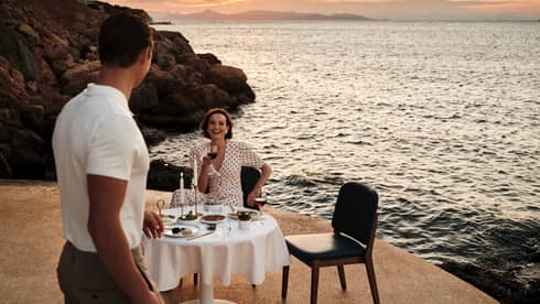 A person sits at an elegant dining table in an outdoor setting, while another person walks towards the table. A rocky shoreline, the setting sun and the ocean are in the background.