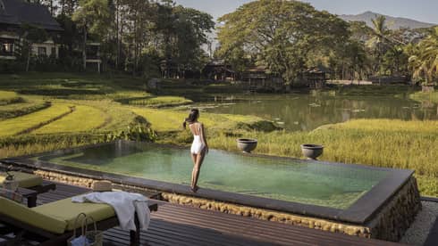 Person standing at the edge of an infinity pool overlooking rice fields, a tranquil pond and forested hills.