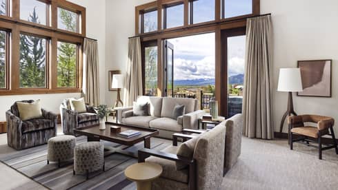 Luxury resort living room with high ceilings, large windows, sofa, armchairs, ottomans, table, floor lamps and doors opening to a mountain-view balcony