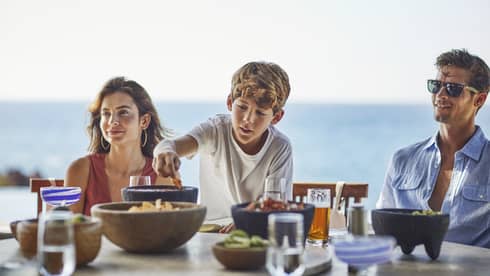 A young boy reaches for a chip bowl on table as a woman and man sit on either side, ocean in the background