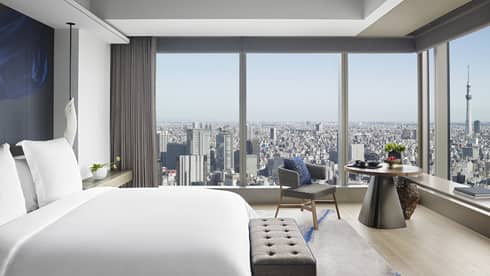 Guest room with king bed, tufted bench, round table with armchair and floor-to-ceiling windows offering city views