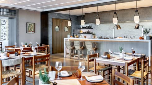 Indoor restaurant with wooden tables and chairs, bar with three wicker seats and pendant lights