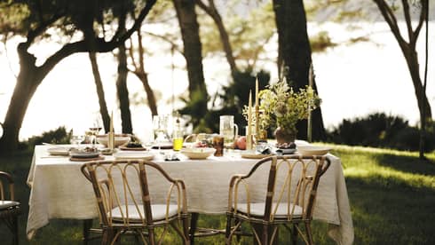 A linen tablecloth adorns an outdoor table set for five with wine, fresh seafood and fruit in an ocean-view forest setting.