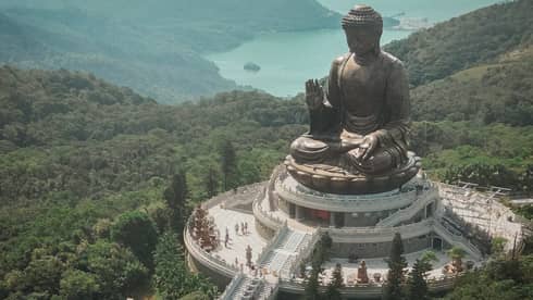 Aerial view of a giant bronze Buddha statue at Po Lin Monastery, surrounded by greenery and mountains. A long staircase leads to the statue on a platform