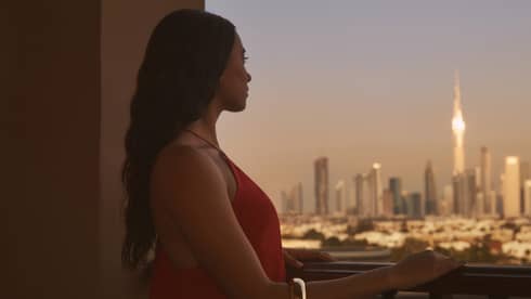 Person with long brown hair standing on balcony with hand resting on railing, looking out to a city at dusk