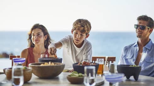A young boy reaches for a chip bowl on table as a woman and man sit on either side, ocean in the background