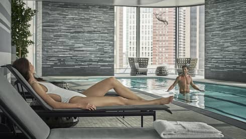 A woman in a white bathing suit and lounge chair looks on at her significant other in the lap pool
