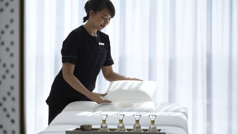 Spa attendant wearing a black uniform fluffs a while pillow on a treatment table covered with all-white linens and a tray filled with four small glass bottles