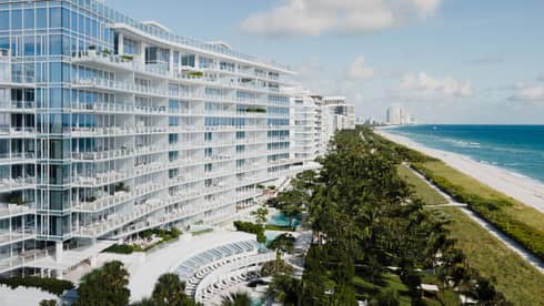 Luxury beachfront hotel with modern glass architecture, lush greenery, a pool area and a view of the ocean and sandy beach under a blue sky