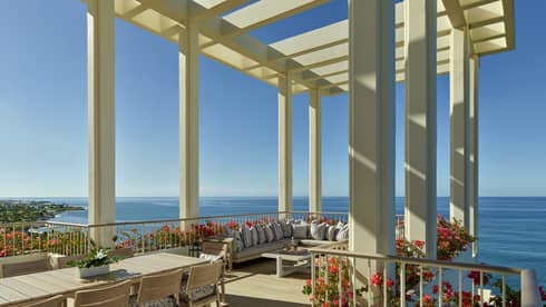 A spacious terrace with a pergola overlooking the ocean, featuring a large dining table, comfortable seating and vibrant flower arrangements under a clear blue sky