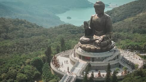 Aerial view of a giant bronze Buddha statue at Po Lin Monastery, surrounded by greenery and mountains. A long staircase leads to the statue on a platform