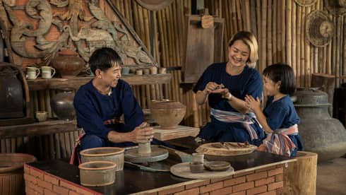 A potter teaching a woman and a child to shape clay in a rustic pottery workshop, surrounded by traditional tools and pottery.